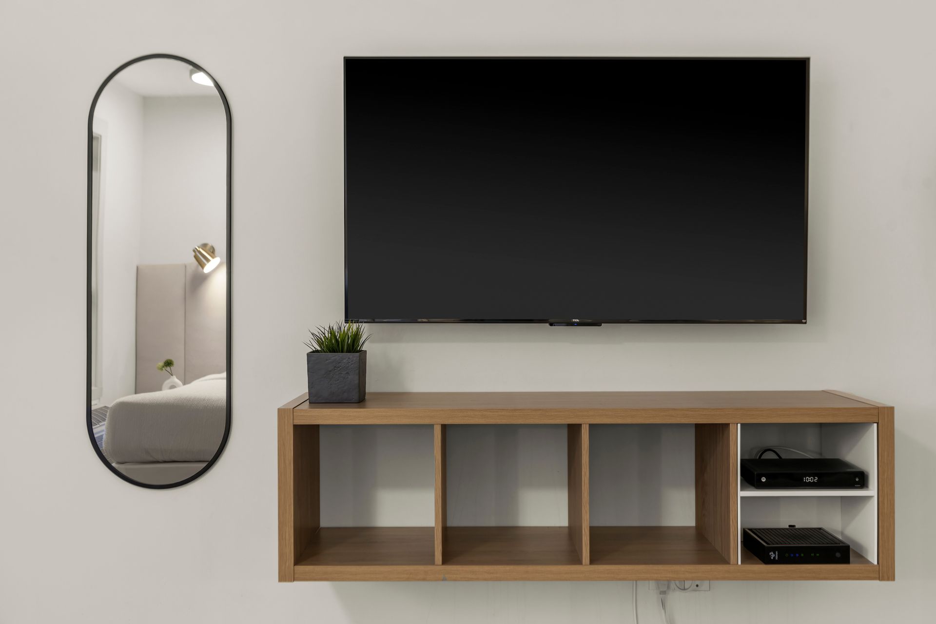A wall-mounted TV above a wooden storage unit with a potted plant and a tall, oval mirror on the left.
