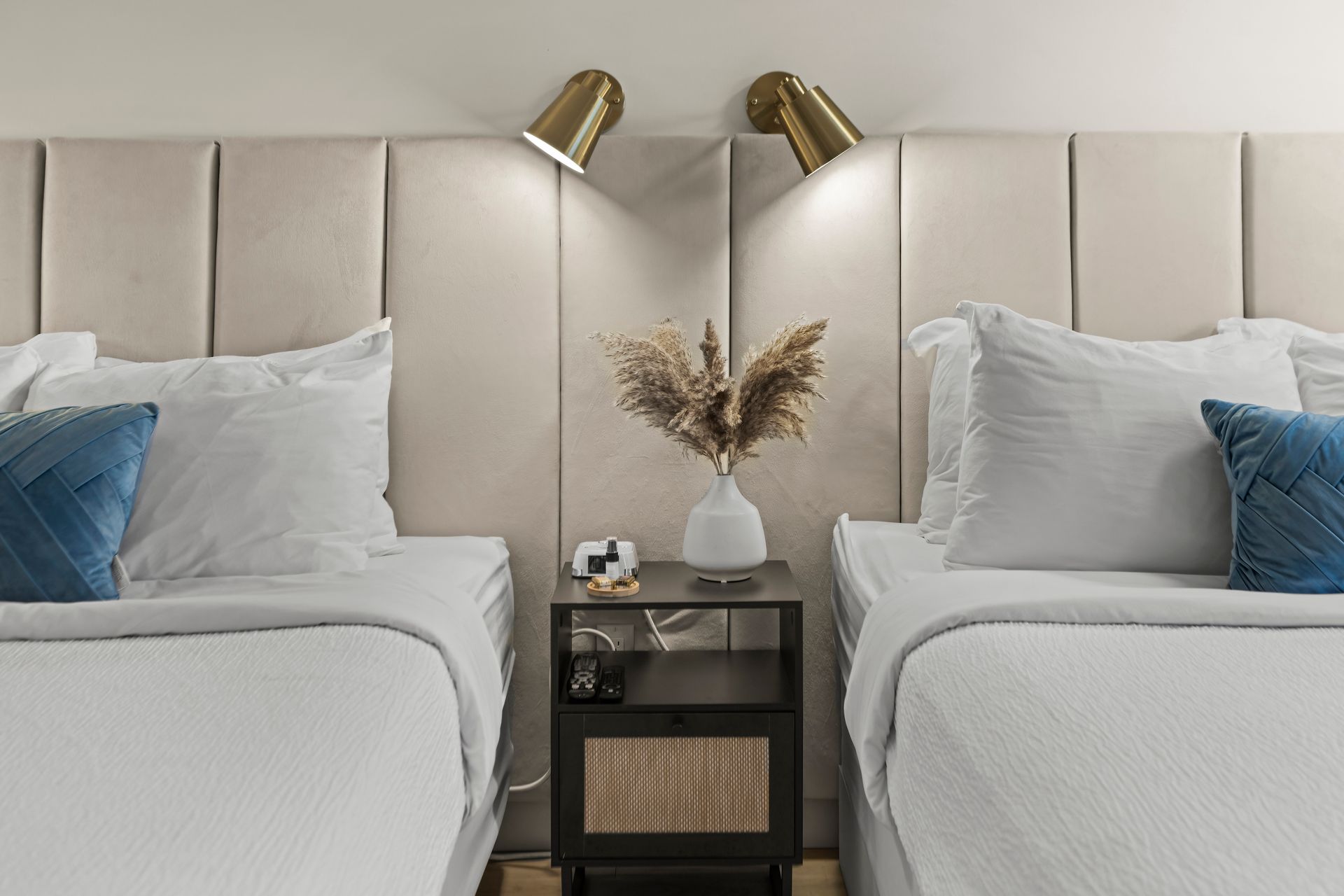Two beds with white linens and blue accent pillows sit on either side of a dark bedside table with a vase of dried flowers.
