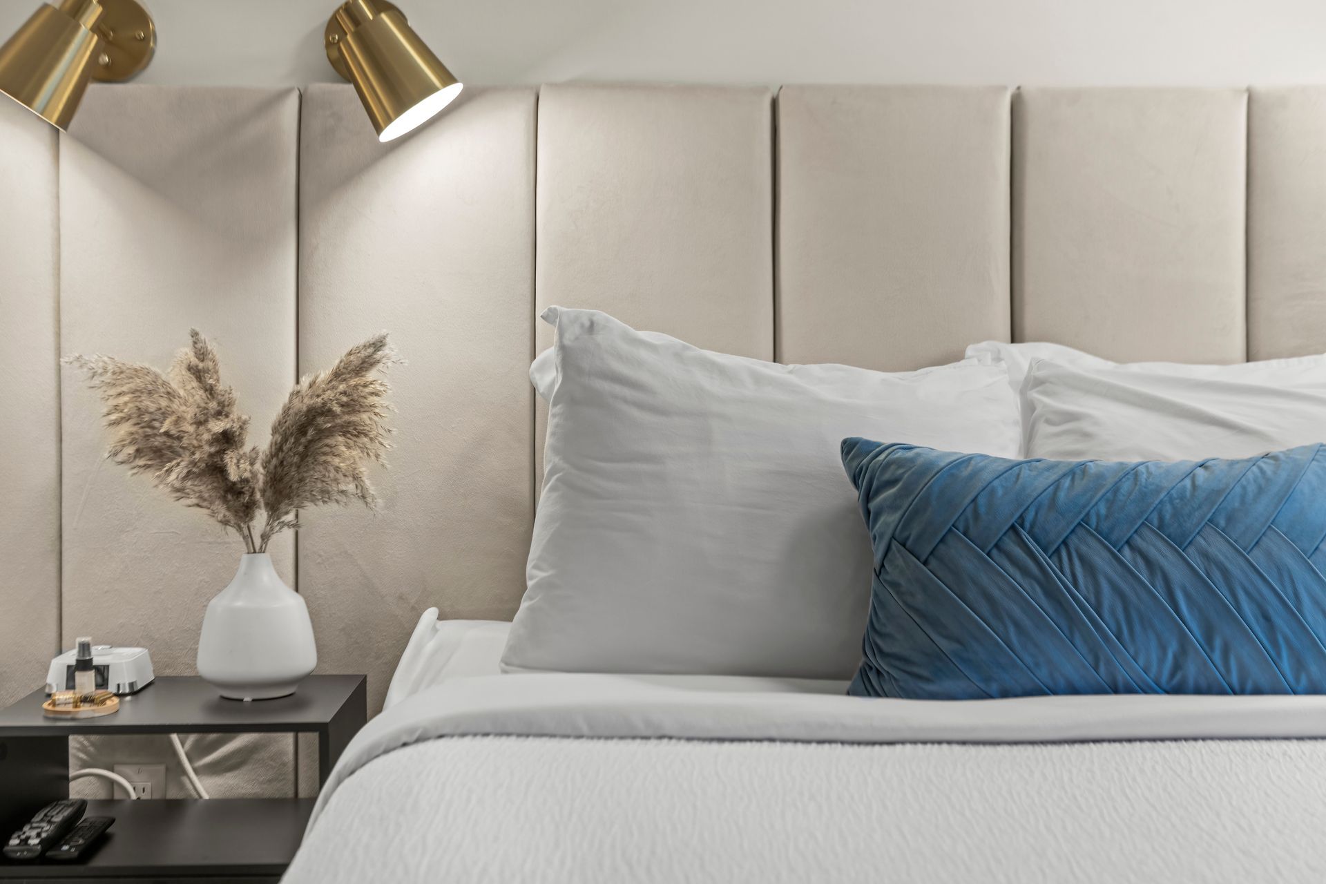 A beige padded headboard, a white pillow, and a blue decorative pillow on a bed with a bedside table and lamp.