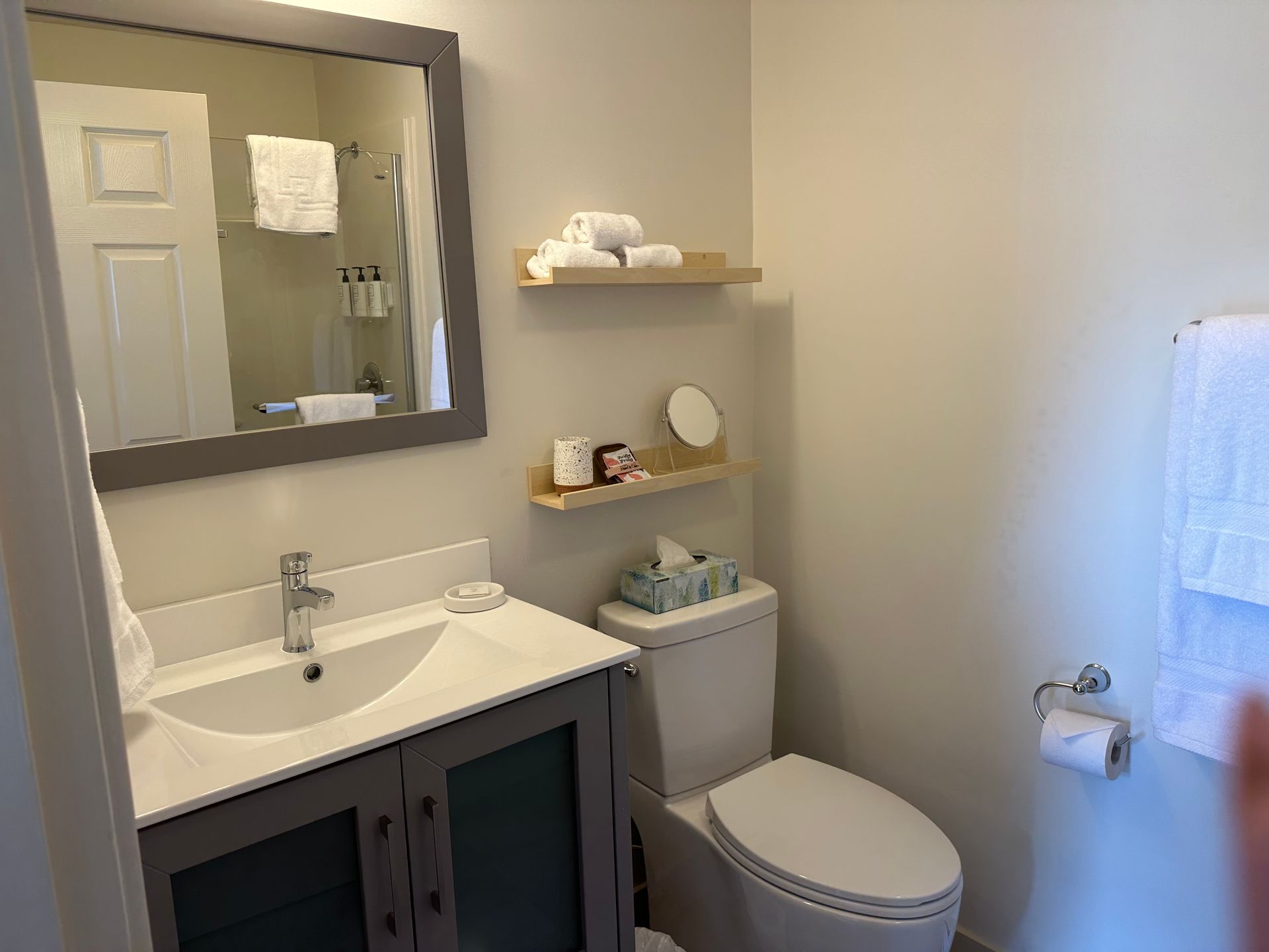 A bathroom featuring a grey vanity with a white sink, a mirror, two floating wood shelves, and a white toilet.