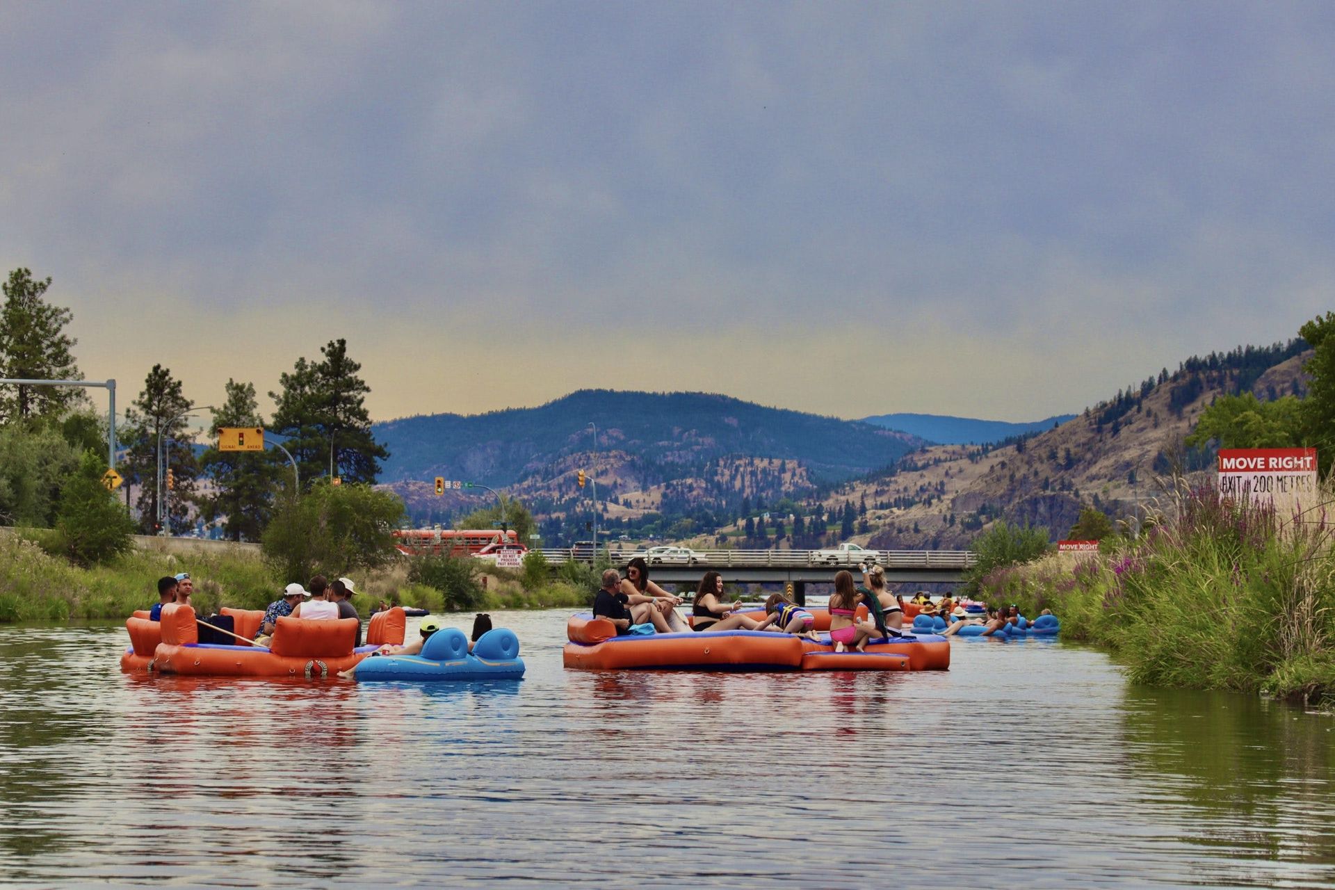 People float on orange and blue inflatable rafts down a river under a cloudy sky, with mountains in the background.
