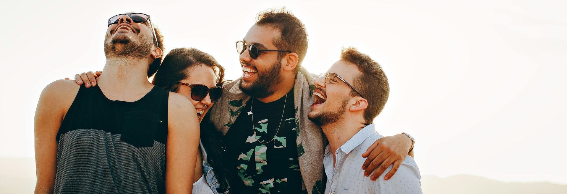 Four friends laughing and embracing outdoors against a bright, hazy sky.