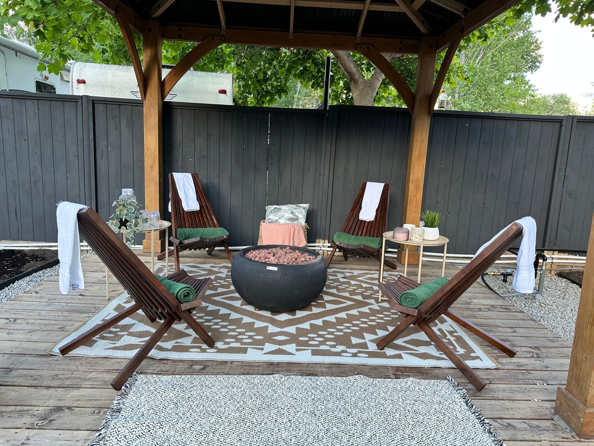 Outdoor seating area with four wooden chairs around a round fire pit on a patterned rug under a wooden gazebo.