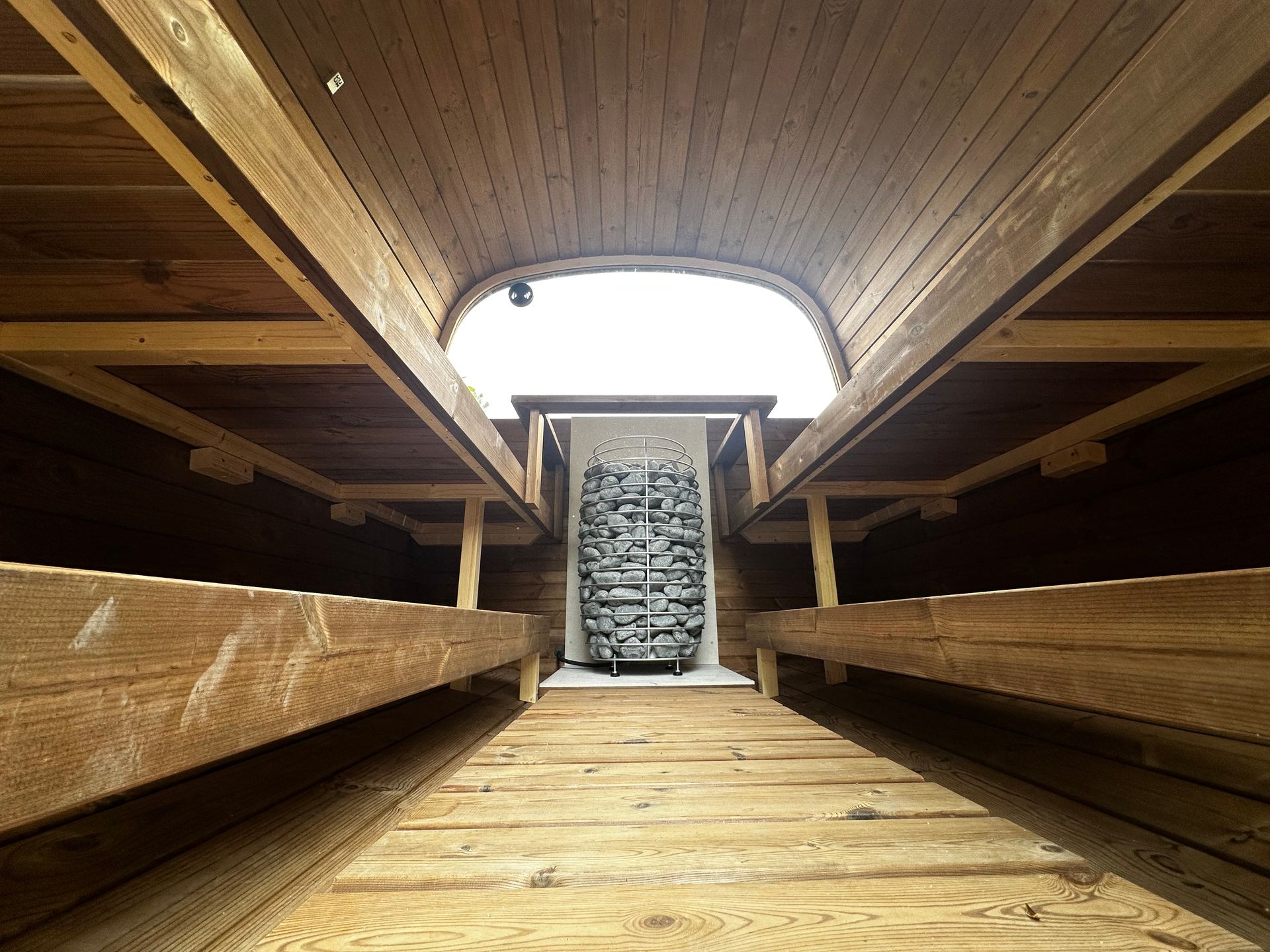 An interior view of a sauna with wooden benches on both sides leading to a central, stone-filled metal heater.