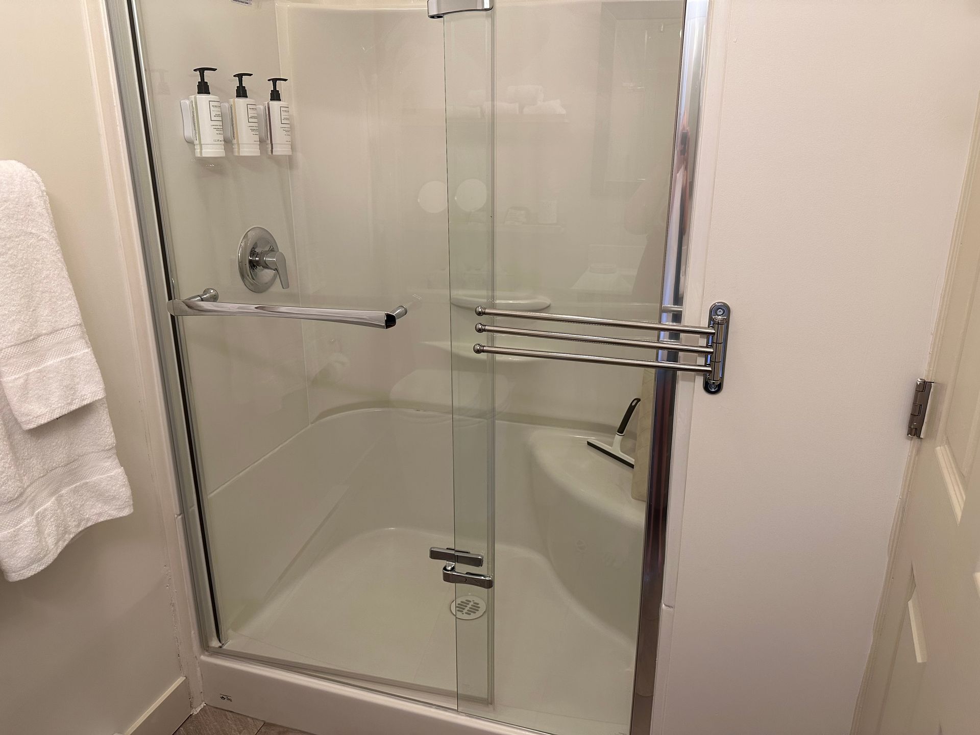 A walk-in shower with white walls, a built-in corner seat, glass doors, and a mounted swing-arm towel rack in a bathroom.