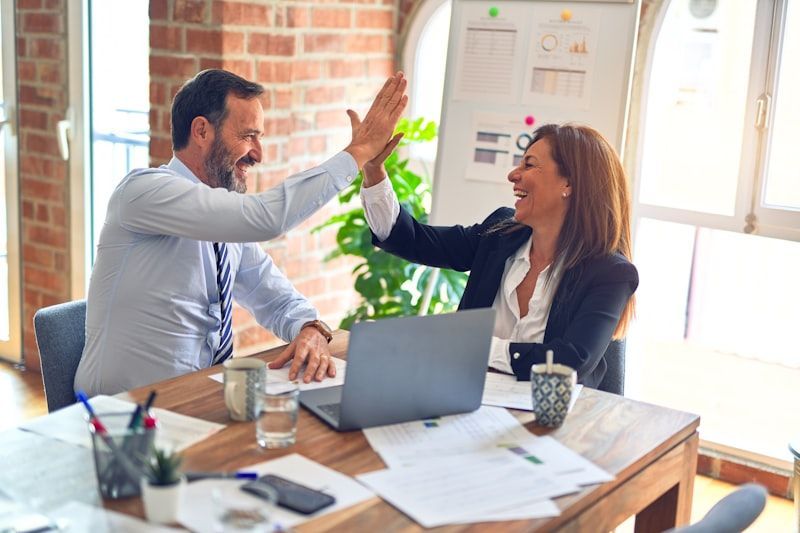 Two professionals in business attire give each other a high-five at an office desk with a laptop and documents.