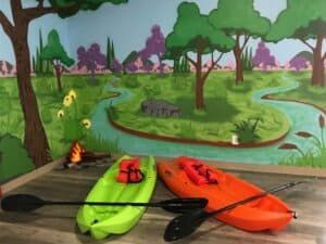 Two kayaks and paddles are sitting on a wooden floor in front of a painting of a river.