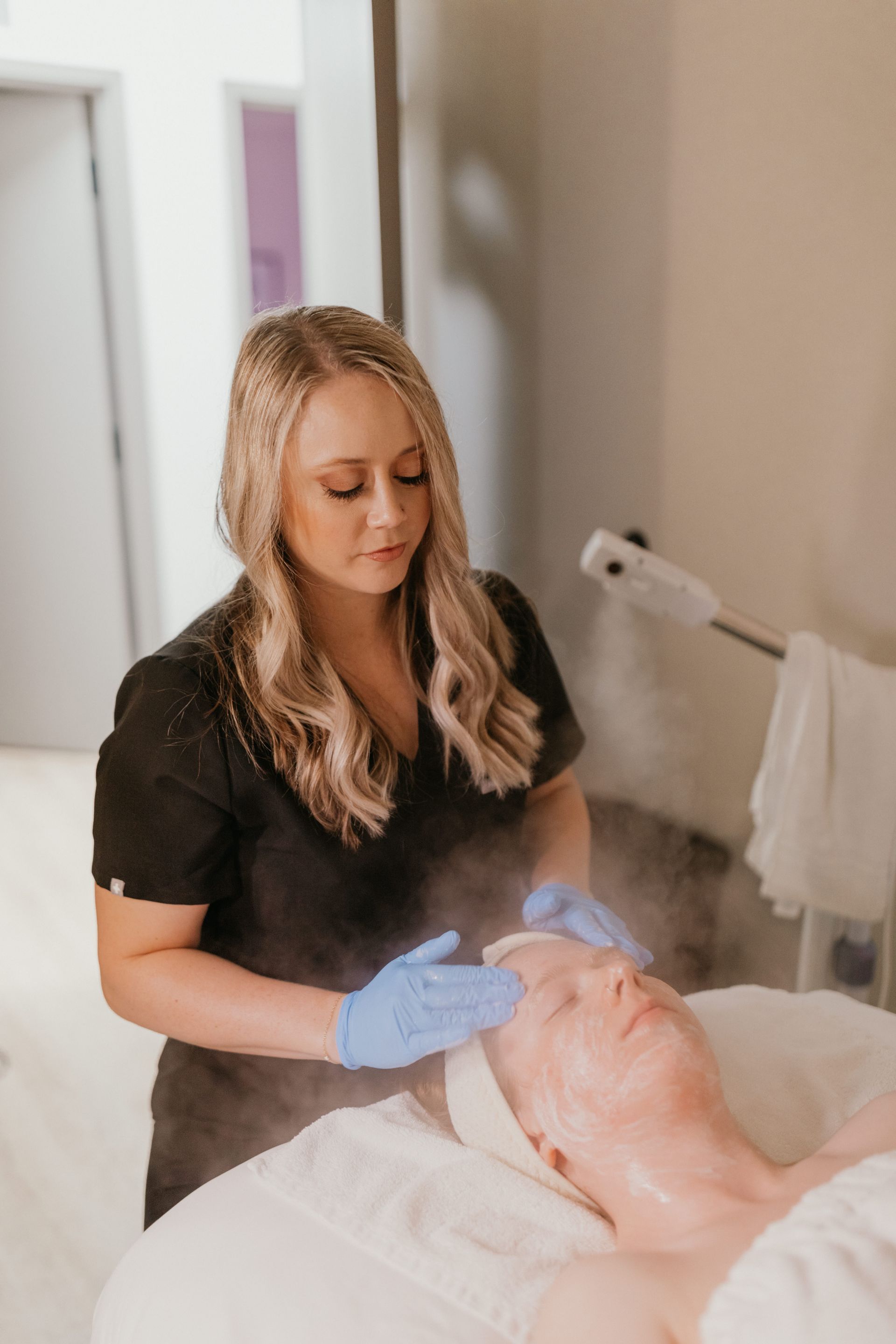 A spa professional in a black uniform and blue gloves performs a facial treatment with steam on a client.