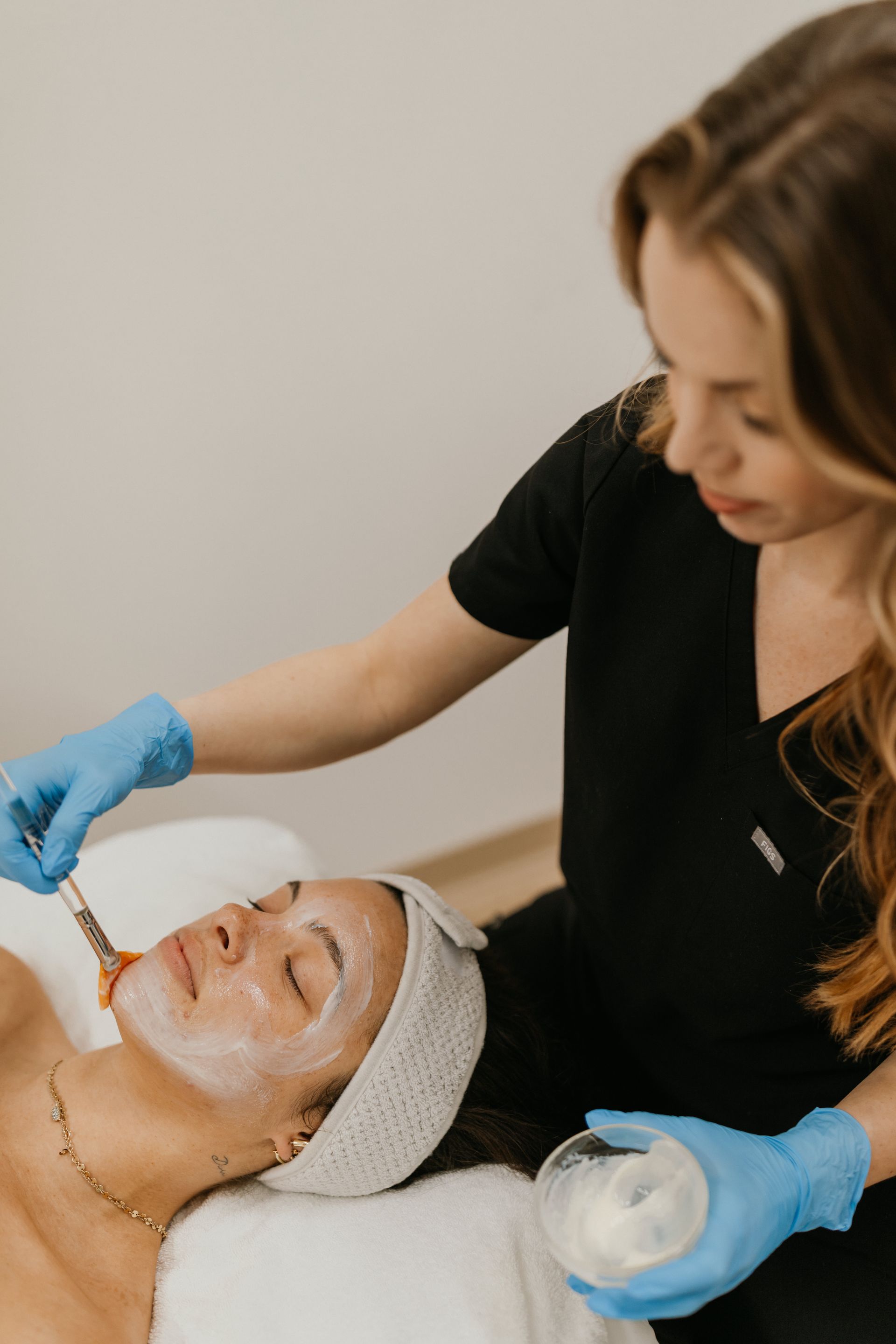 Facial treatment: esthetician applying product to client's face with a brush. Spa setting, both wearing blue gloves.