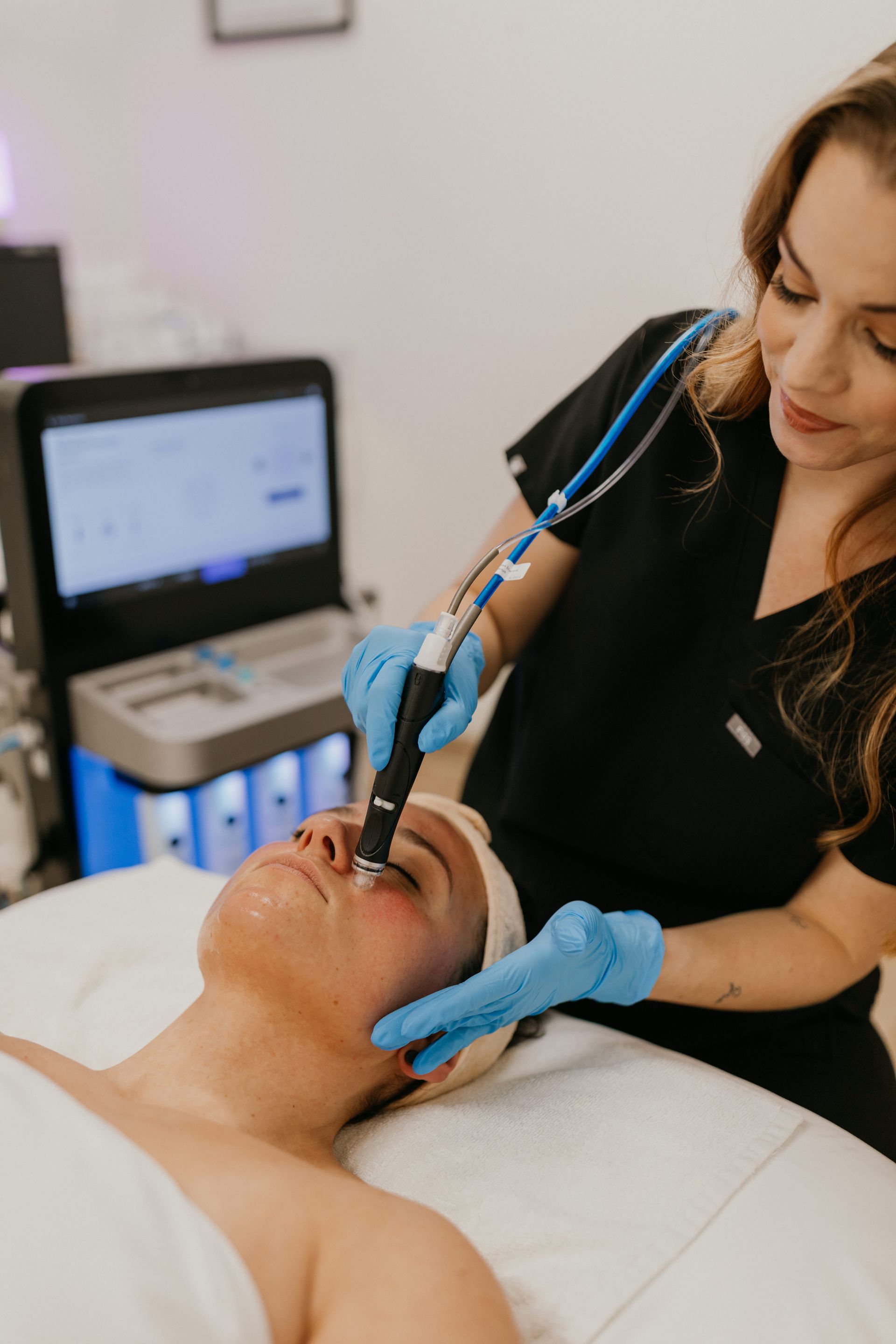 A clinician in a black uniform performs a facial treatment on a client using a handheld wand in a medical spa setting.