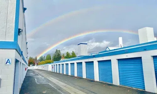 Rainbow over blue storage units beside a wet alley on an overcast day