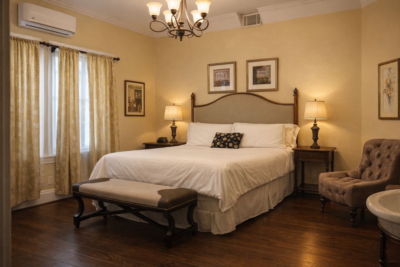 Bedroom with a bed, bench, armchair, lamps, window, and decorative wall art. Yellow walls, hardwood floors.