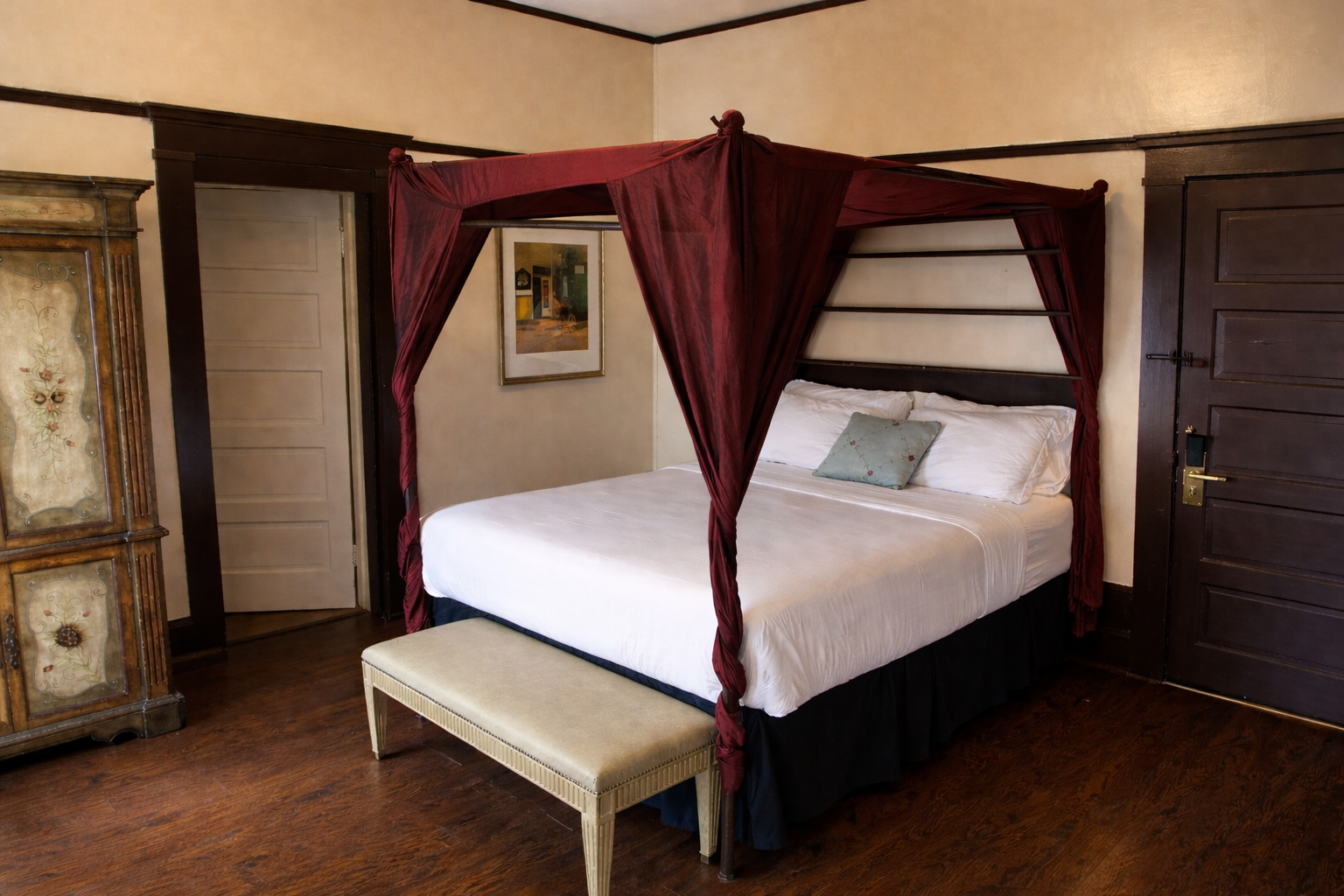 Bedroom with canopy bed, white linens, dark wood trim, light wood floor, and a bench.