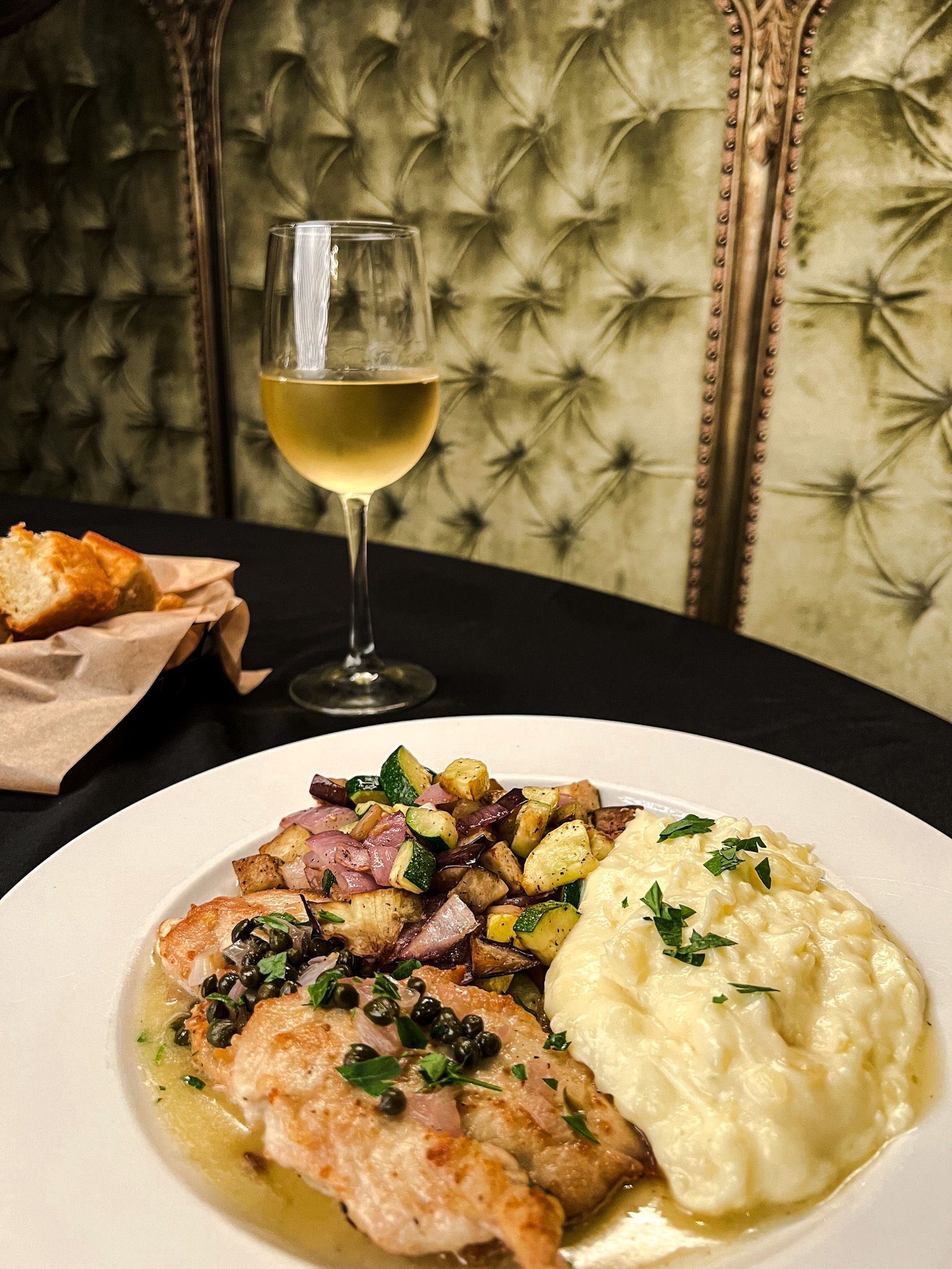 Plate of food with fish, potatoes, and wine on a table with a green textured wall background.