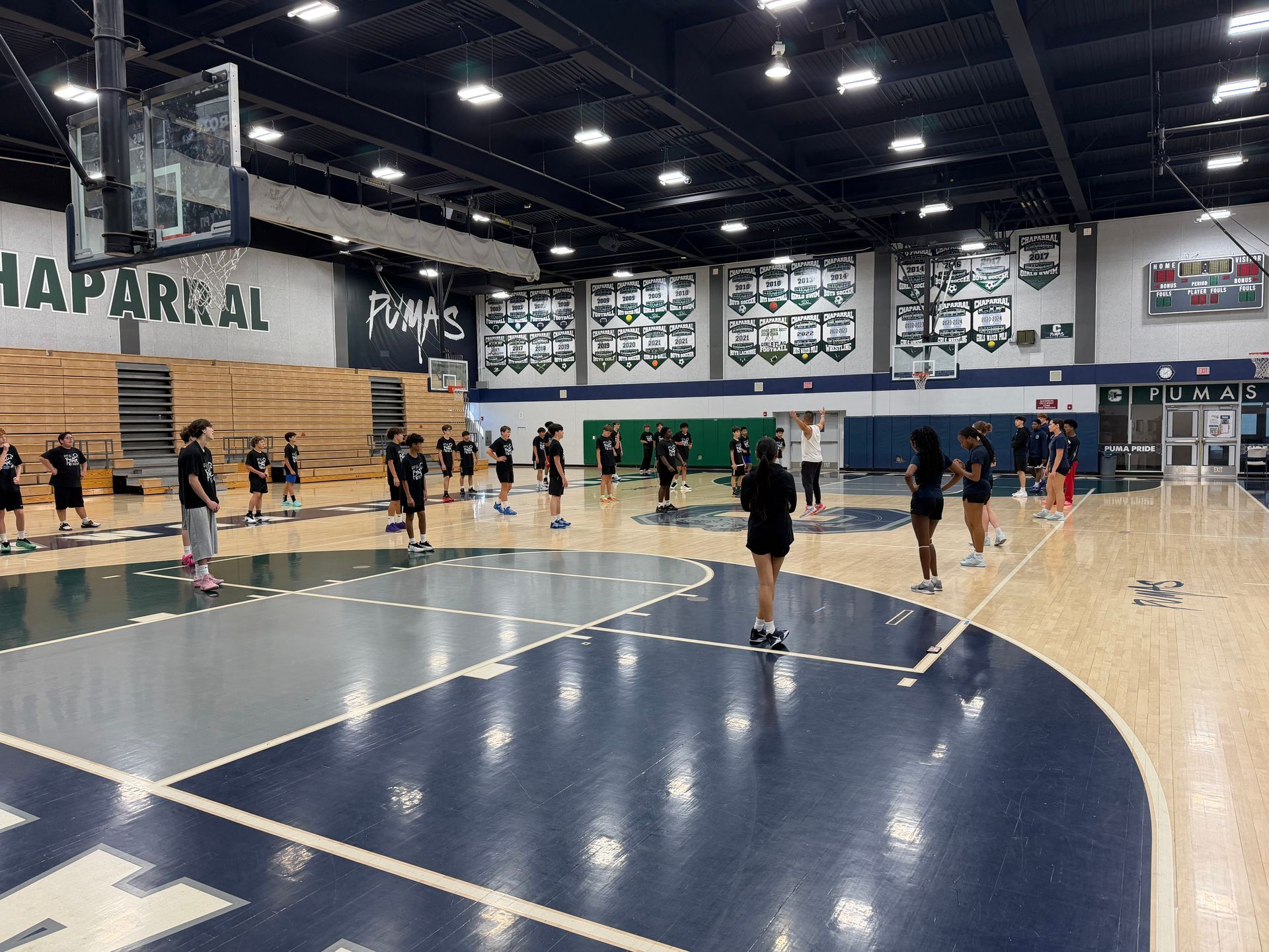 Athletes perform drills on a dark blue and wood-toned basketball court inside a brightly lit indoor sports facility.