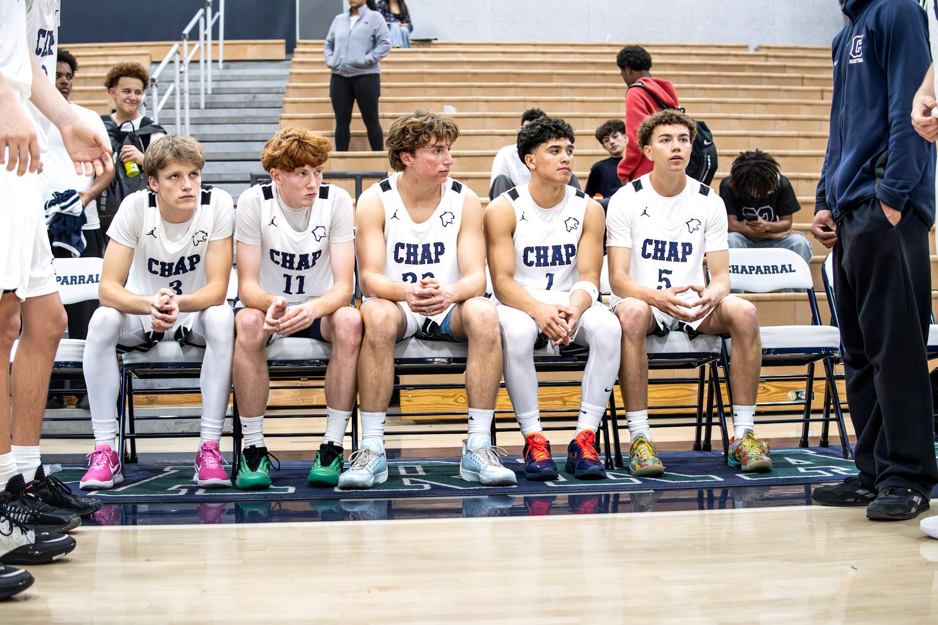 Five basketball players in white uniforms sit on a bench in a gymnasium, watching a game.