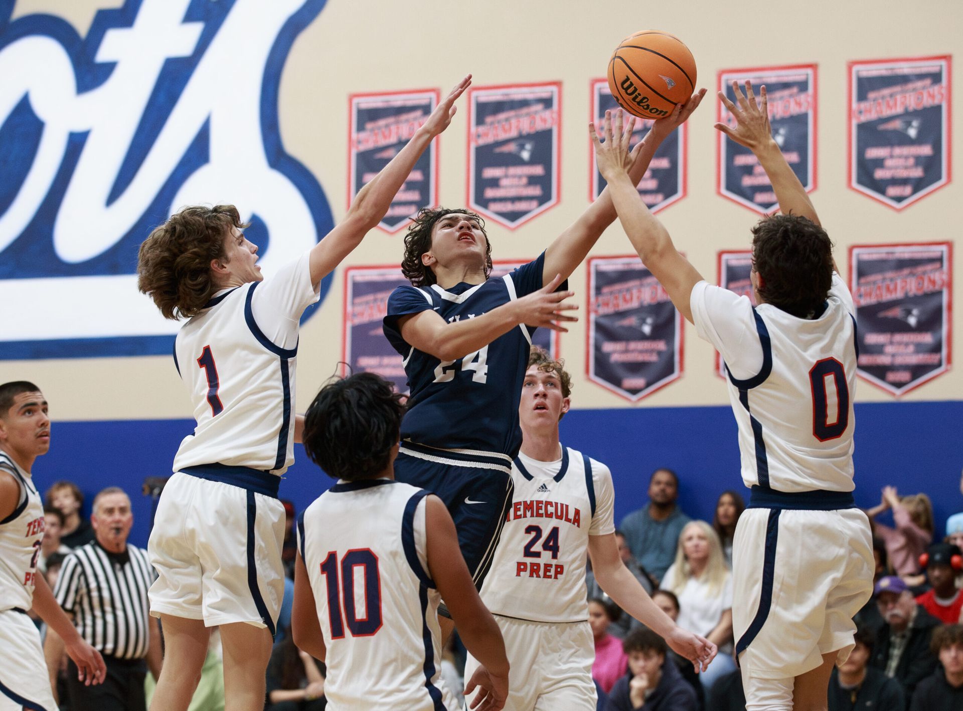 A basketball player in a dark blue uniform jumps to shoot as two opponents in white jerseys defend in a gymnasium.