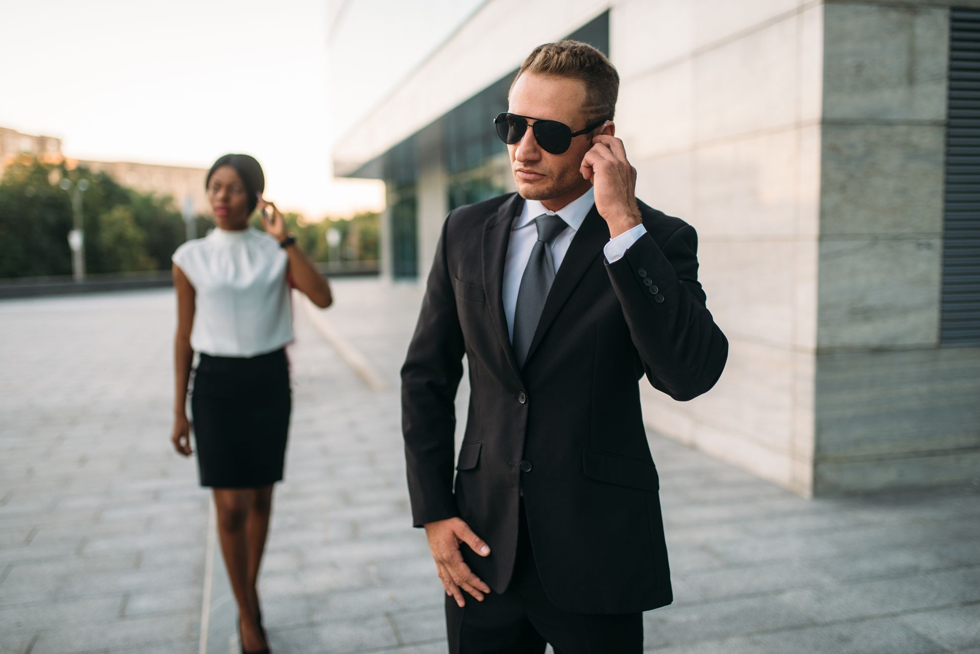 Two businesspeople in suits walking outside a modern office building, one adjusting sunglasses.