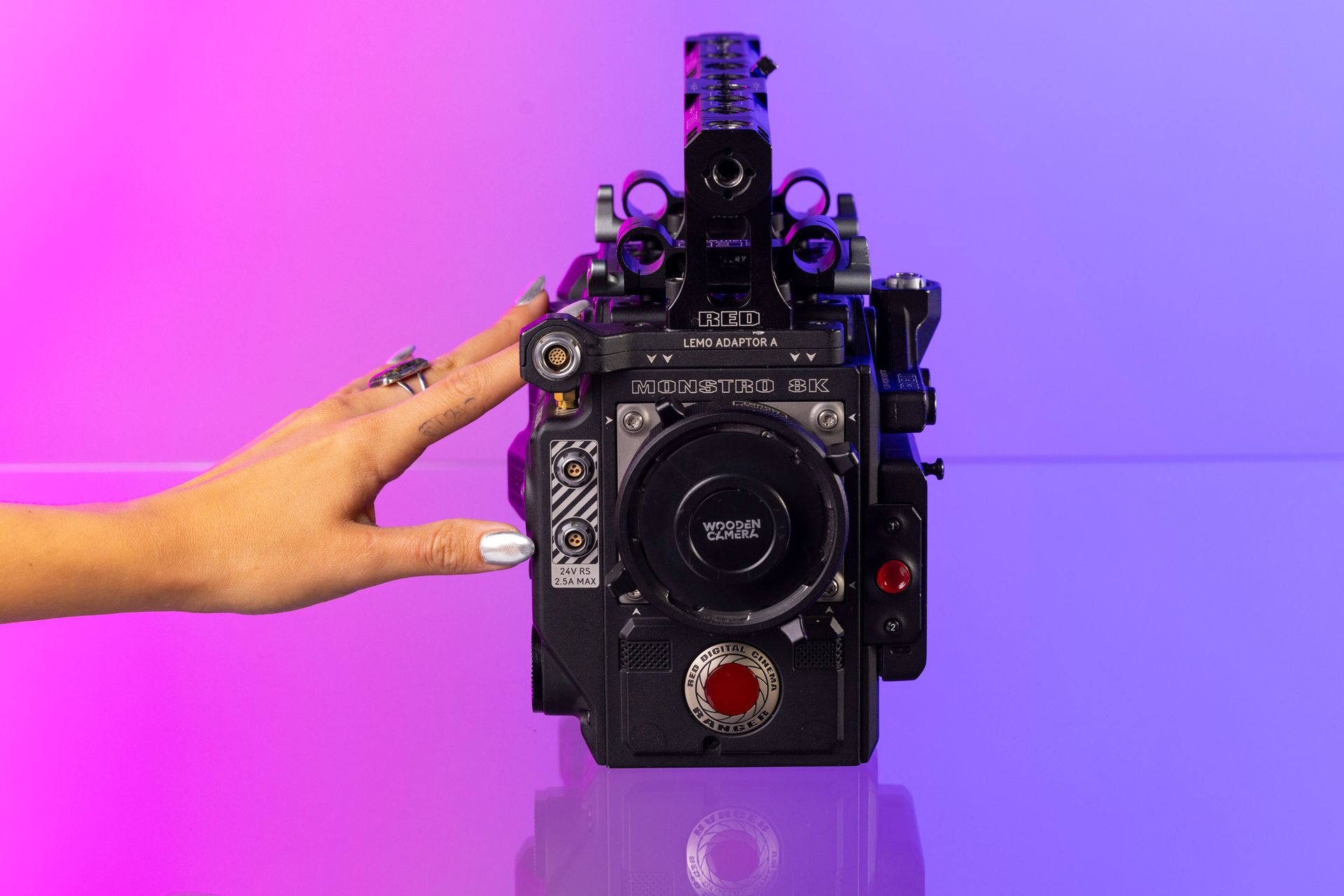 Une main touchant une grande caméra vidéo noire sur un fond dégradé rose et violet.