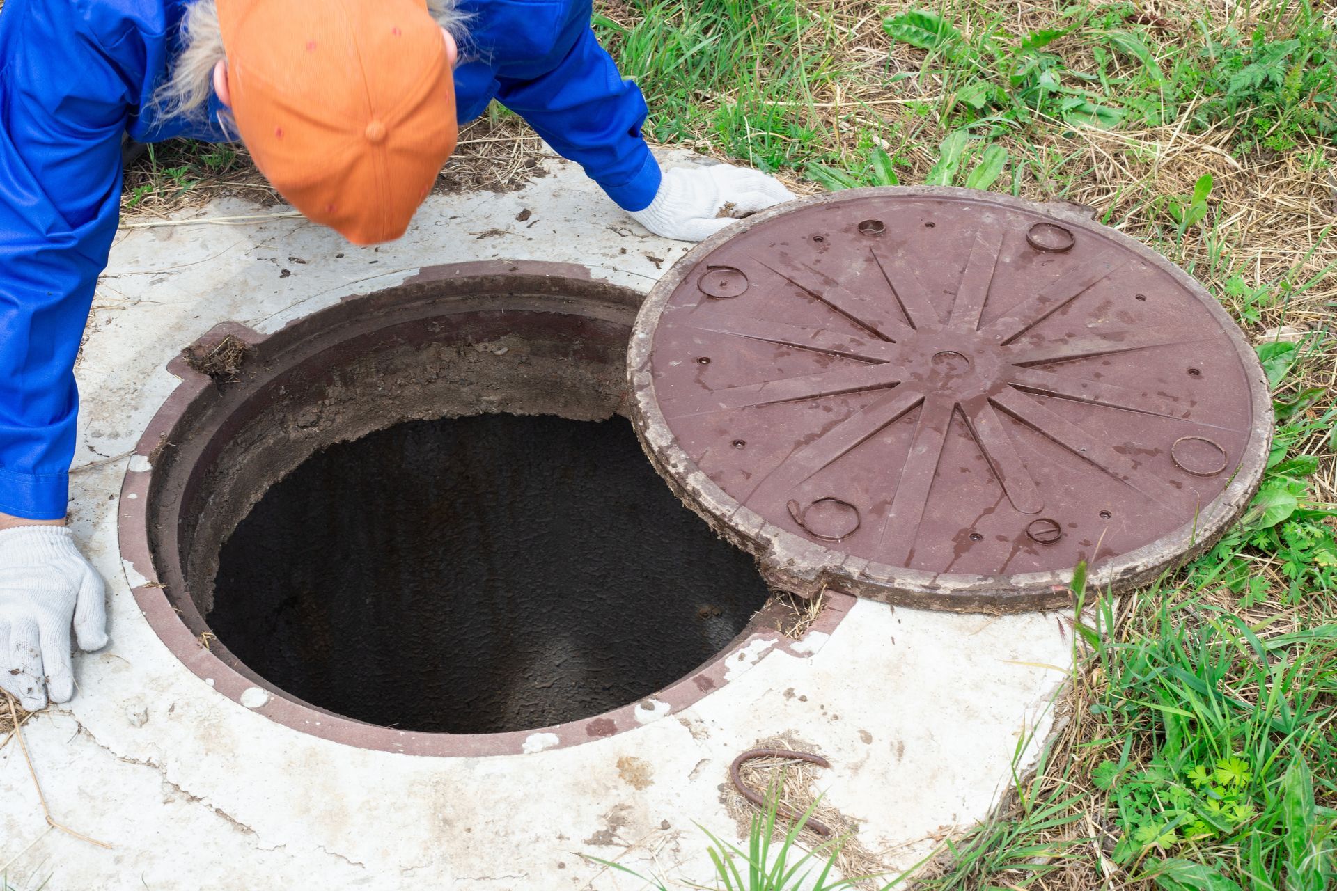 Person in blue suit and orange hat inspects open manhole cover in grassy area.