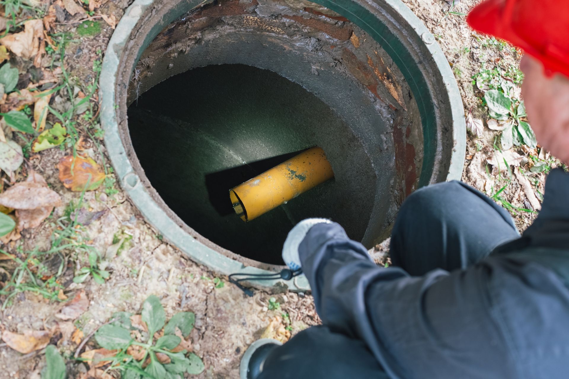 Person inspecting an open septic tank, wearing a red hat, observing inside with a pipe visible.
