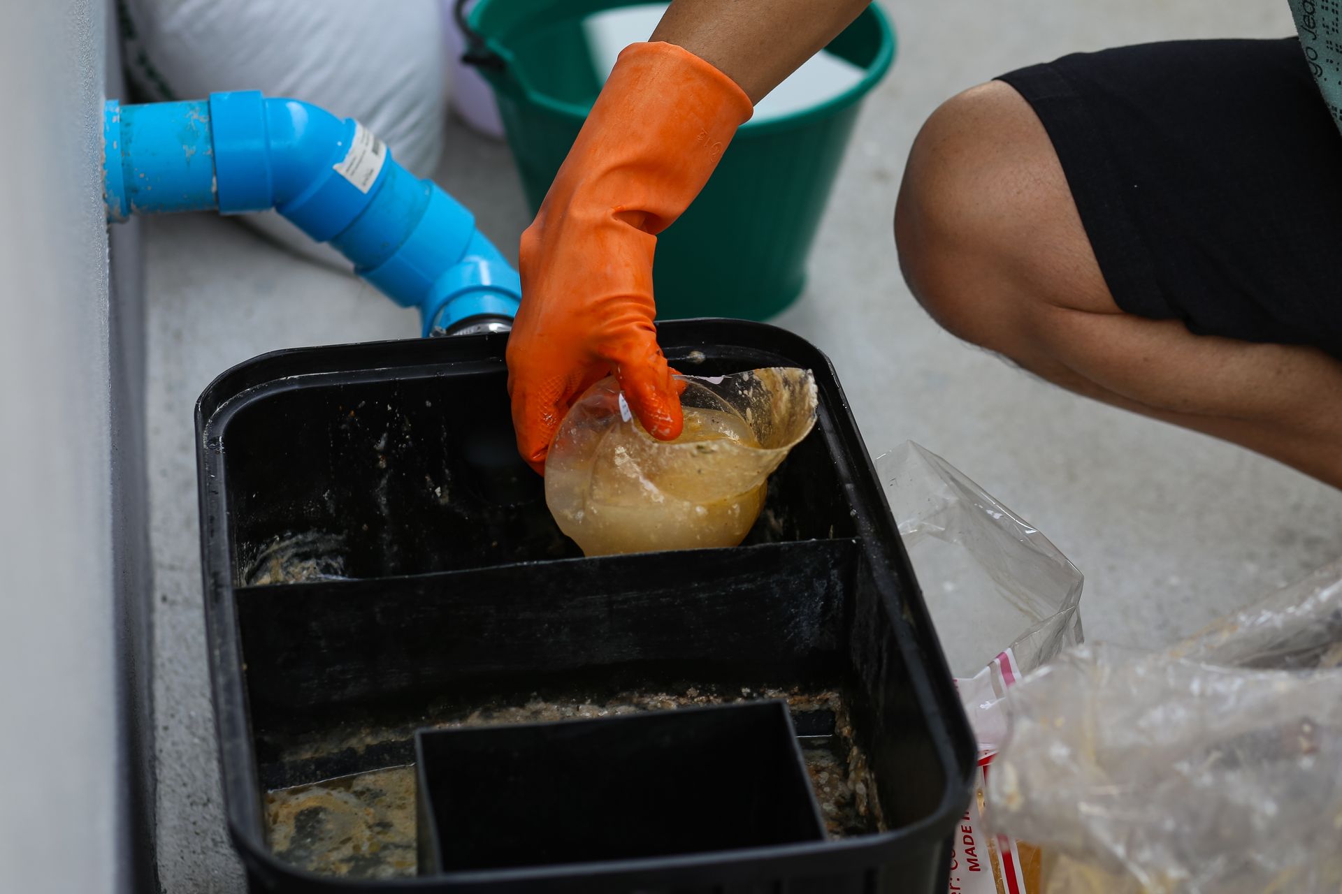 Person in orange gloves removing debris from a black filter box, with blue pipe and green bucket nearby.