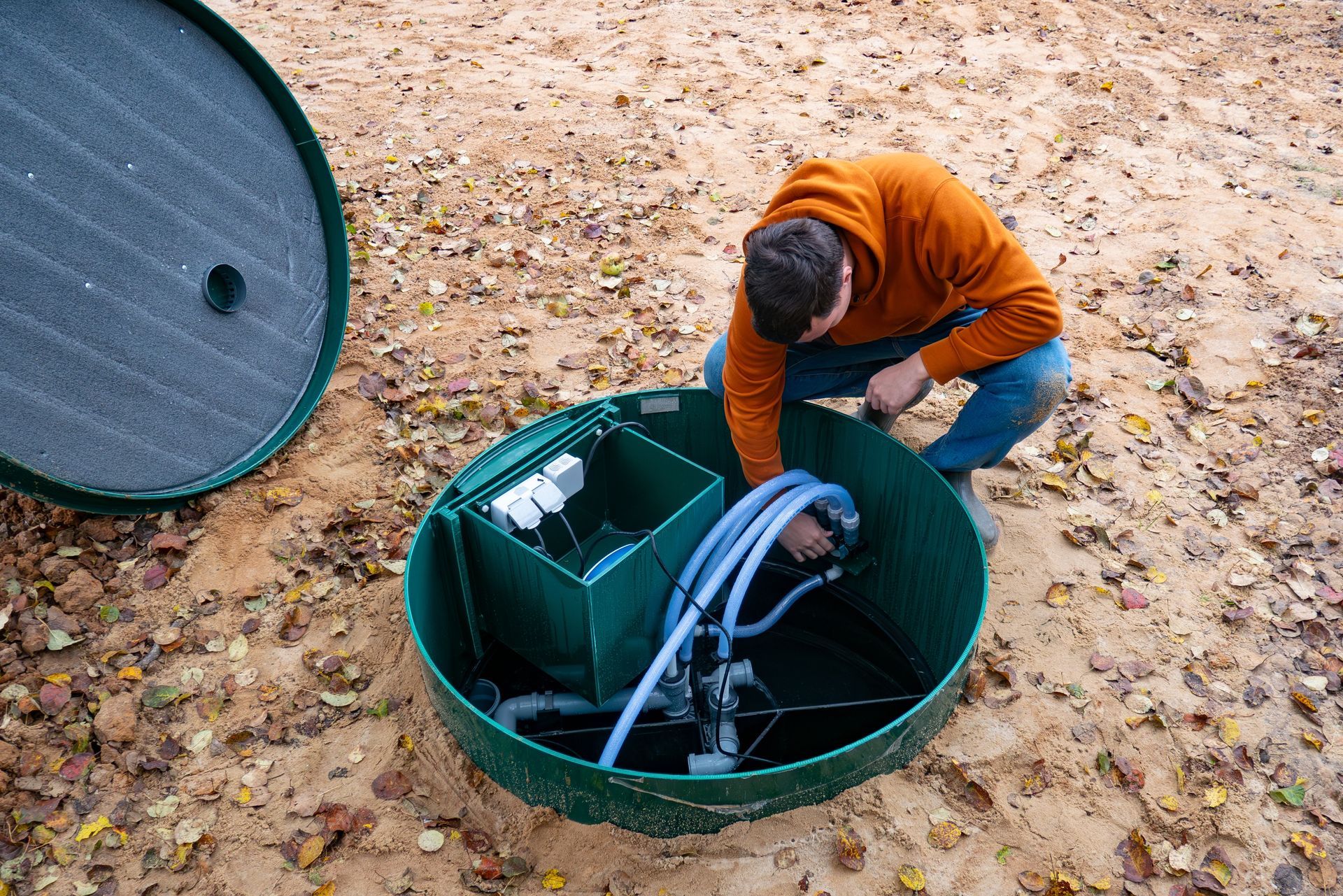 Man inspecting a septic tank outdoors; green tank, blue hoses, orange hoodie.