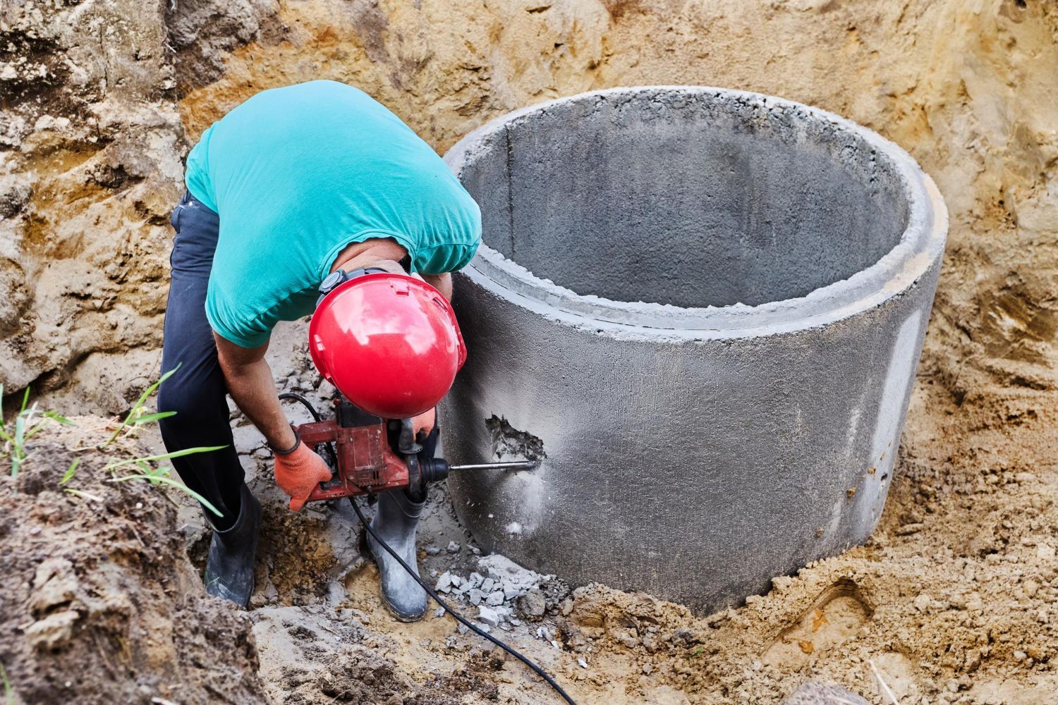 Worker in a red hard hat drilling into a concrete manhole at a construction site