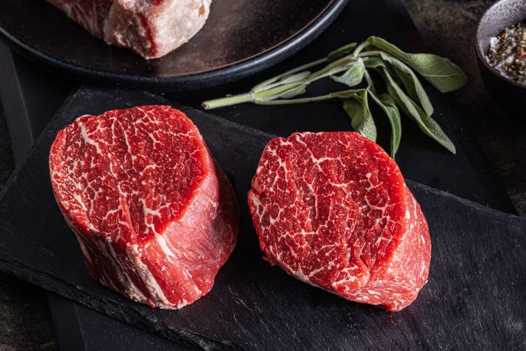 Two steaks are sitting on a black cutting board next to a plate of meat.