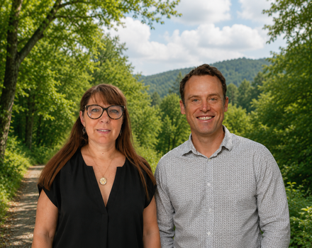 Deux personnes se tiennent à l'extérieur, devant des arbres et des collines verdoyantes, souriant toutes deux à l'objectif.