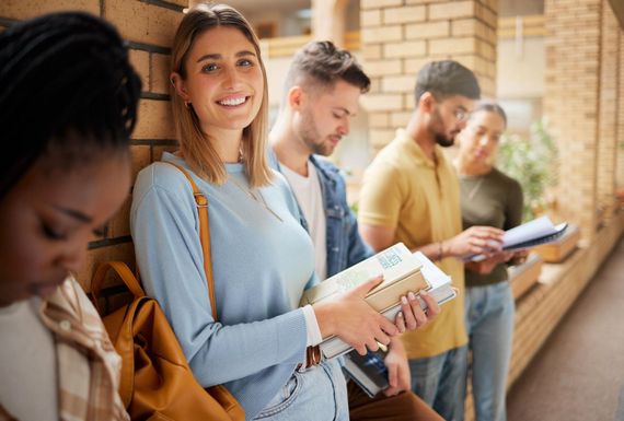 Des étudiants font la queue devant un bâtiment en briques, tenant des livres et des cahiers.