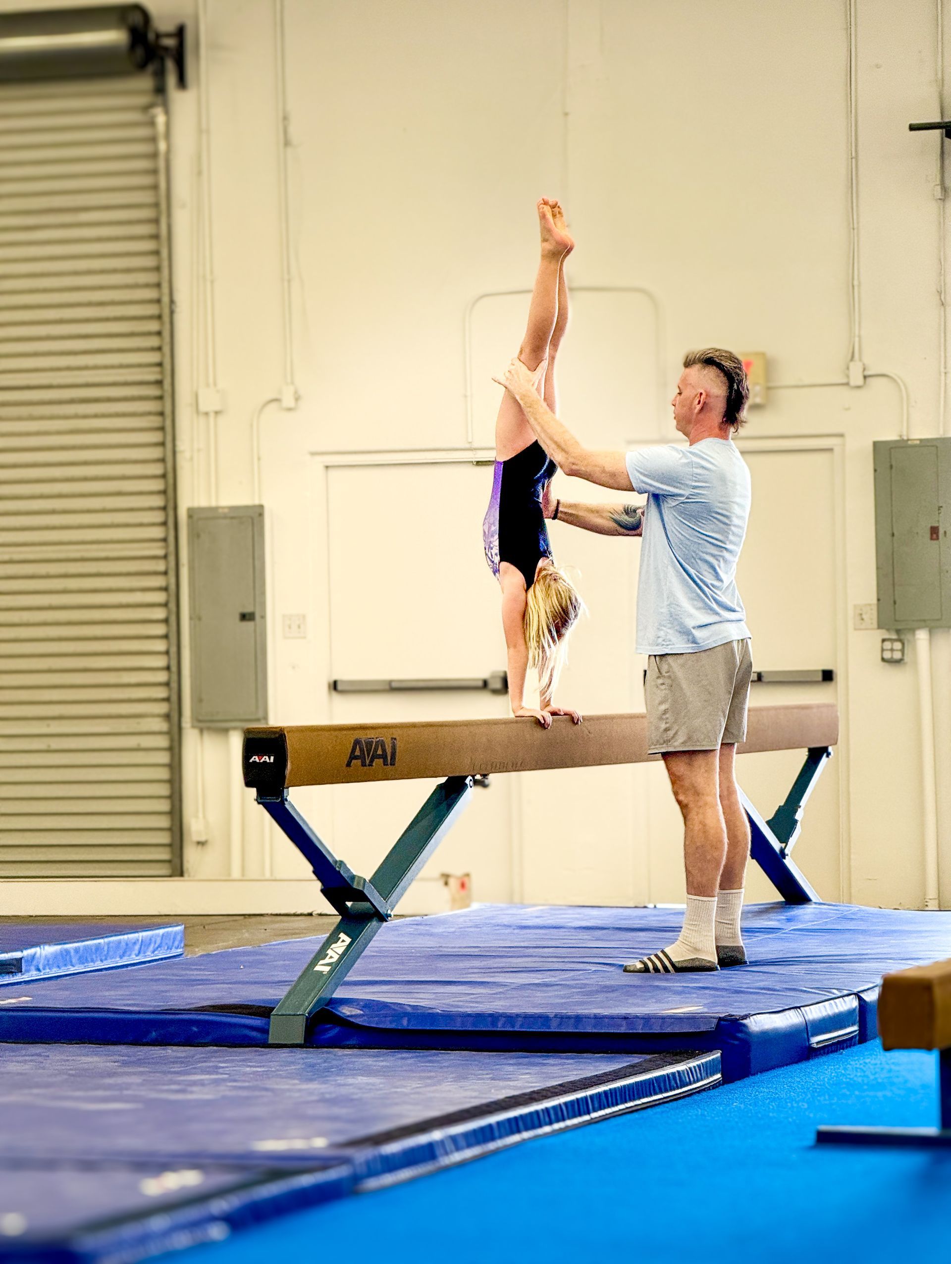 Gymnast on balance beam with coach assisting a handstand. Indoor gym setting.