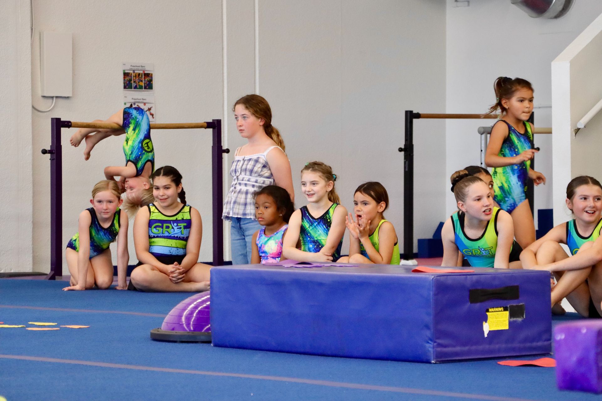 Gymnast on balance beam, instructor assisting. Gym with colorful cones, other gymnasts.