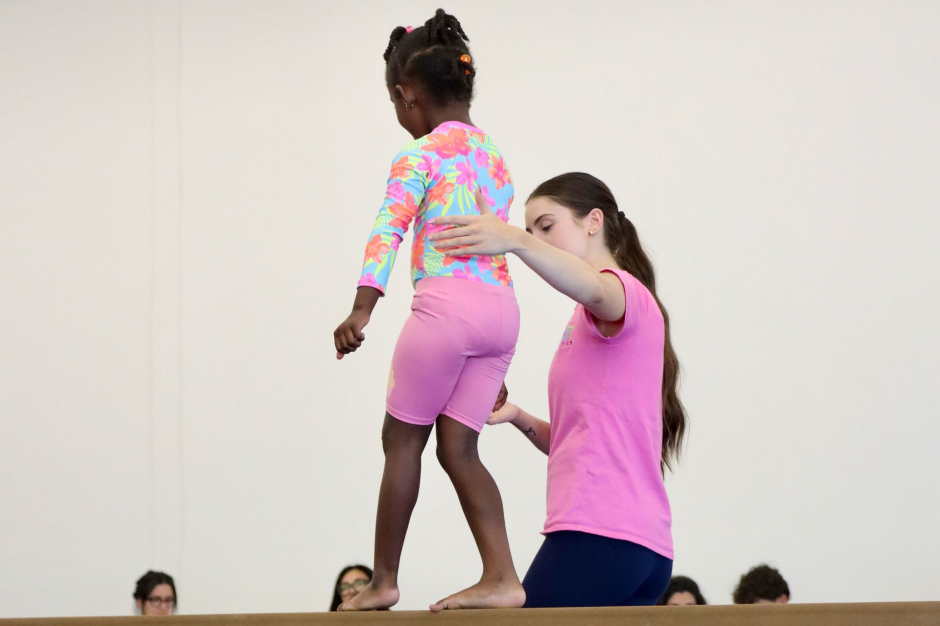 Gymnast on balance beam, instructor assisting. Gym with colorful cones, other gymnasts.