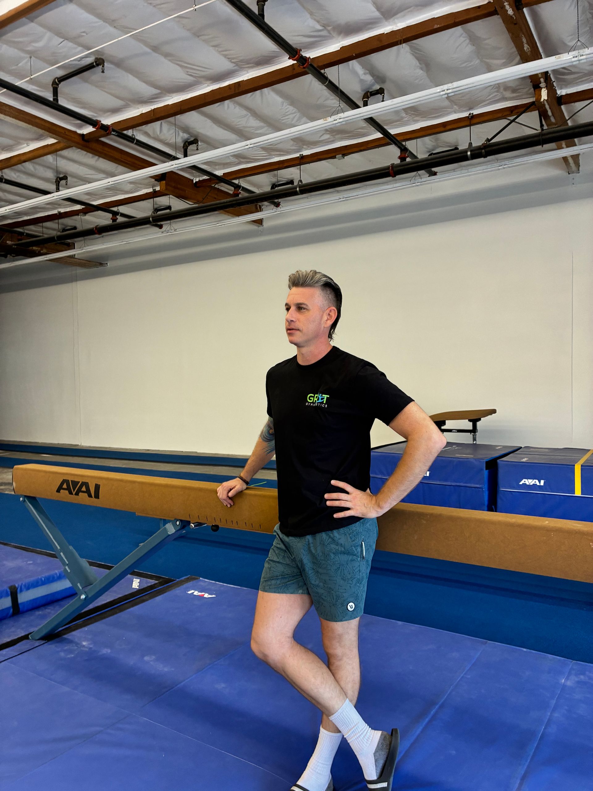 A person stands leaning against a balance beam in a gym, wearing a black t-shirt, patterned shorts, and white socks.