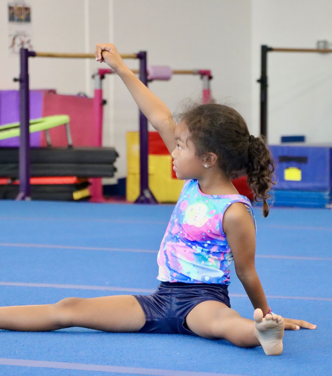 Girl on balance beam, another girl spots her. Gym setting, red beam, blue mat.