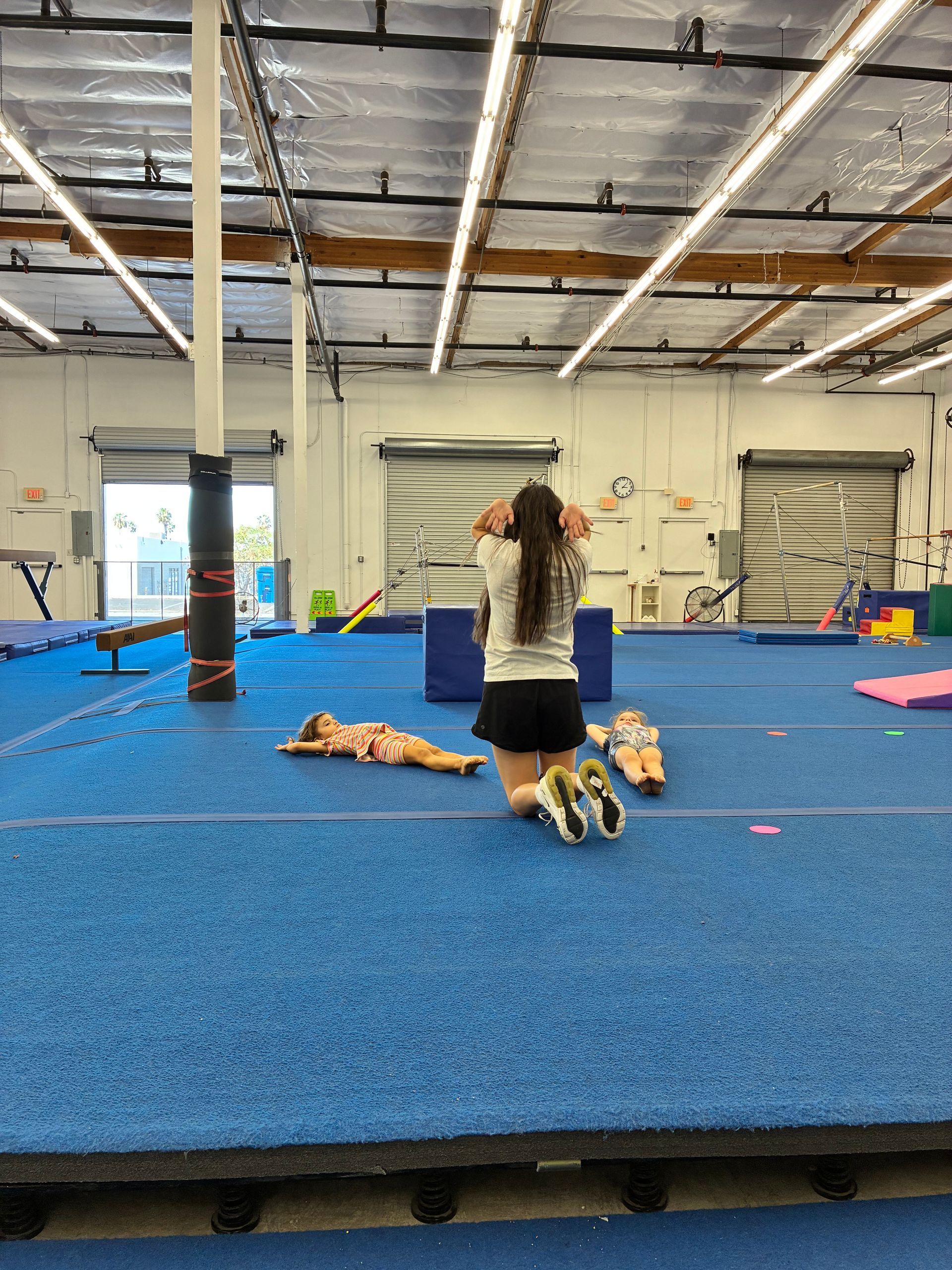 Gymnastics instructor on knees, observing two children on the blue floor of a gym.