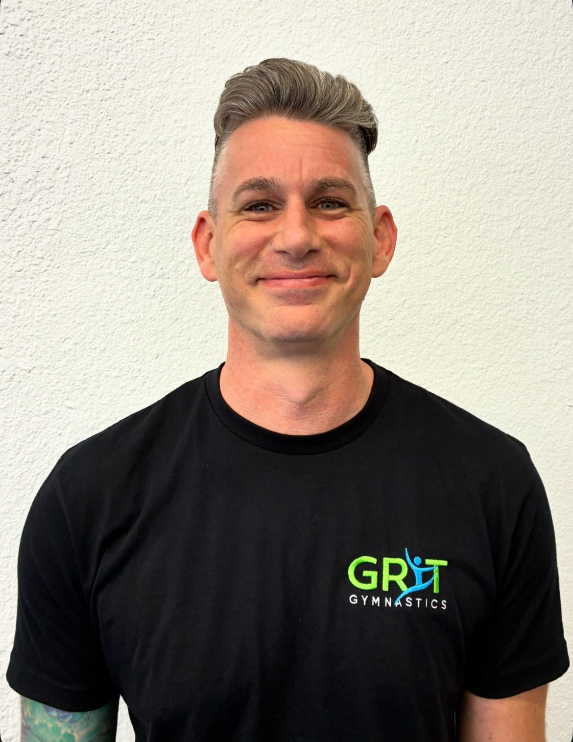Man wearing a black t-shirt with “GRIT GYMNASTICS” logo. He is smiling in front of a white wall.