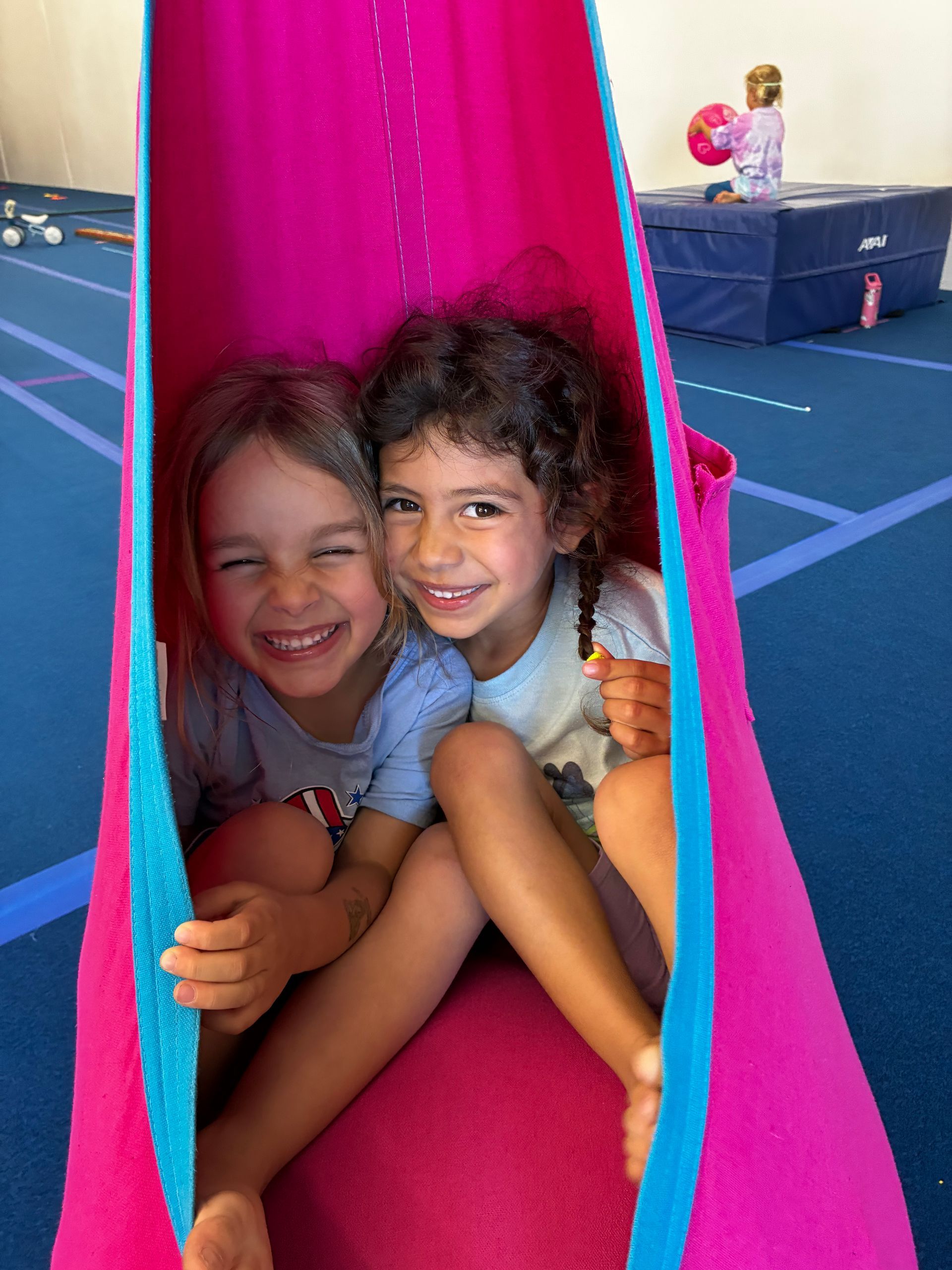 Two children smiling inside a bright pink play tunnel on a gym floor