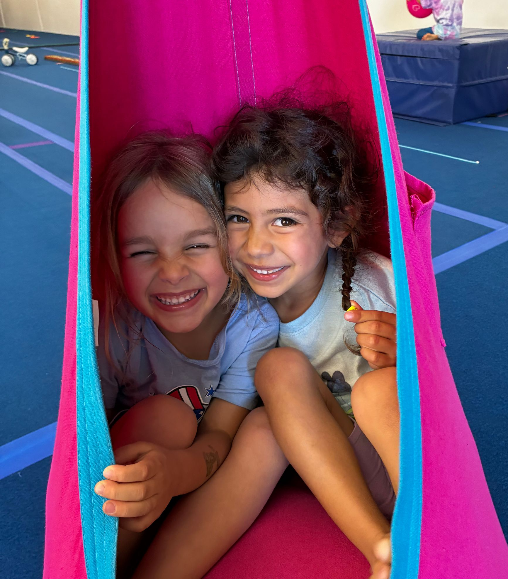 Gymnast on balance beam, instructor assisting. Gym with colorful cones, other gymnasts.