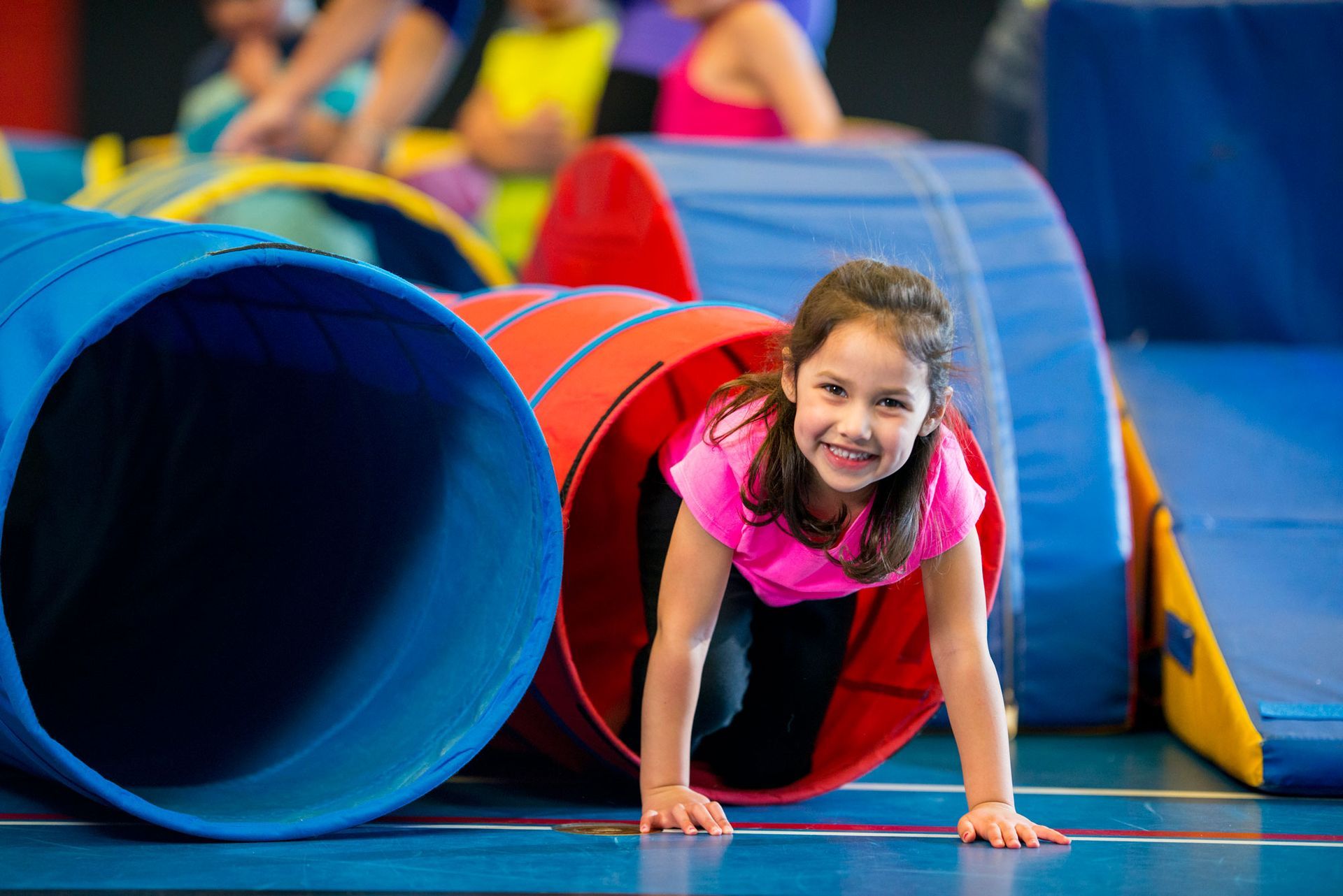 Girl crawling through a red tunnel, smiling, in a colorful indoor play area.