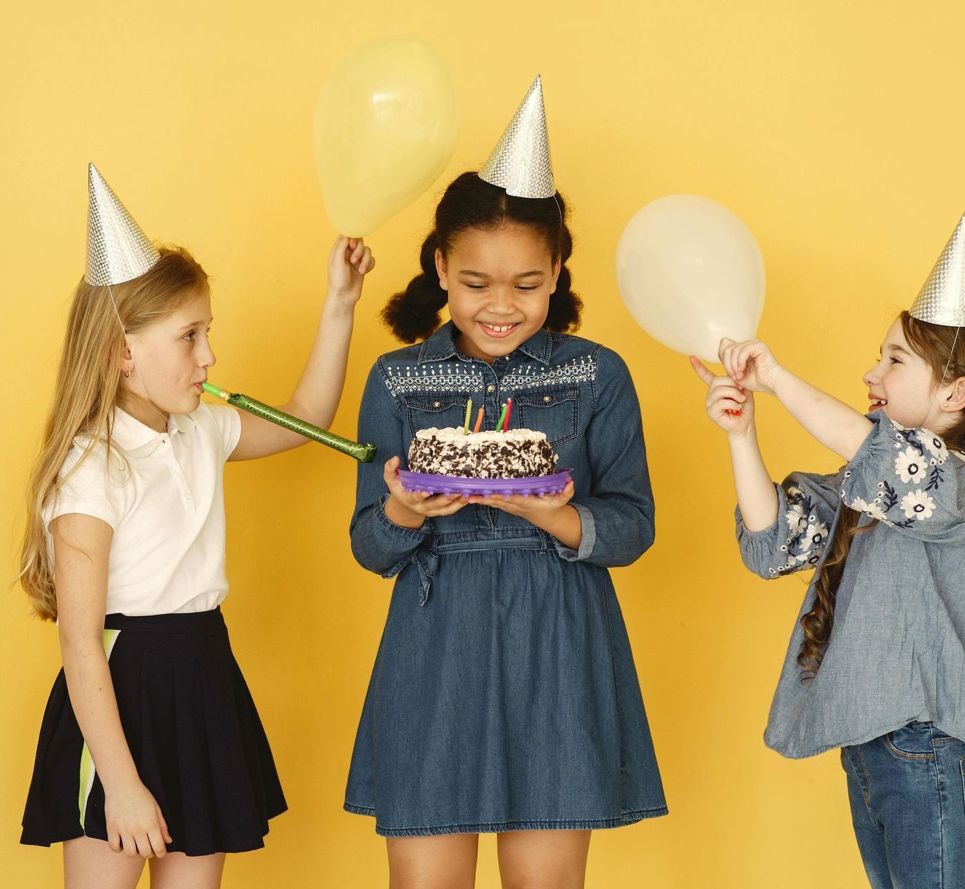 Three girls in party hats celebrating a birthday with a cake and balloons against a yellow background.