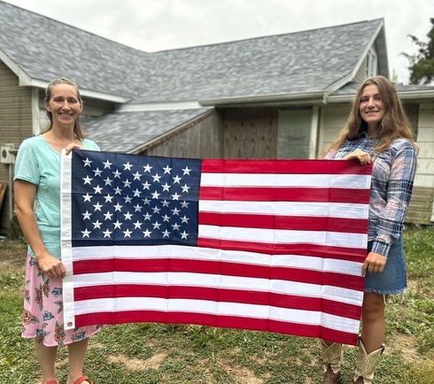 Two people holding a large American flag in front of a house and grassy yard