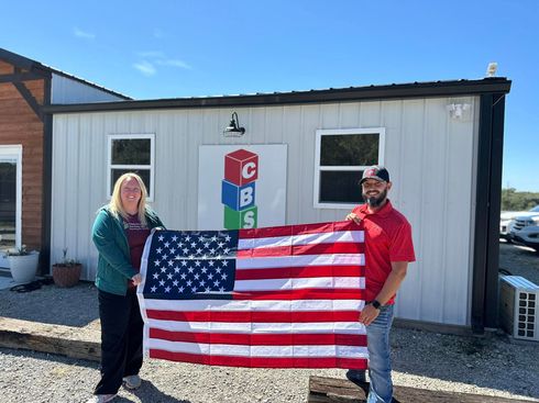 Two people hold an American flag in front of a small building under a blue sky.