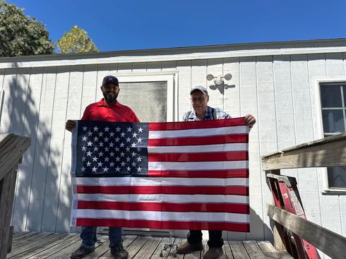 Two people holding a large American flag in front of a white house on a sunny day