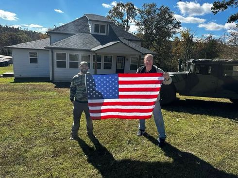 Two people holding a U.S. flag on a lawn in front of a house on a sunny day