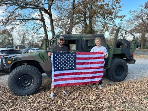 Two people stand by a military jeep holding an American flag in a leaf-covered outdoor setting.