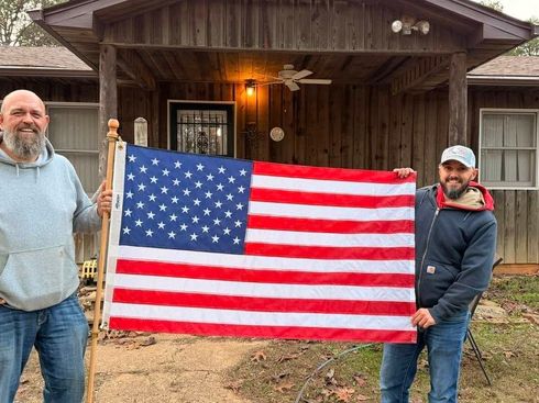 Two men standing outside a cabin holding a large American flag