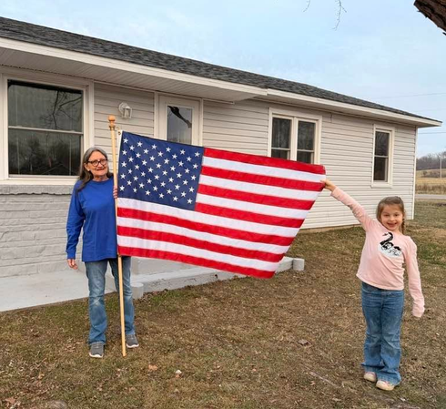 Two people holding a large American flag in front of a house on a cloudy day