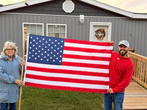 Two people holding a large American flag in front of a gray house.
